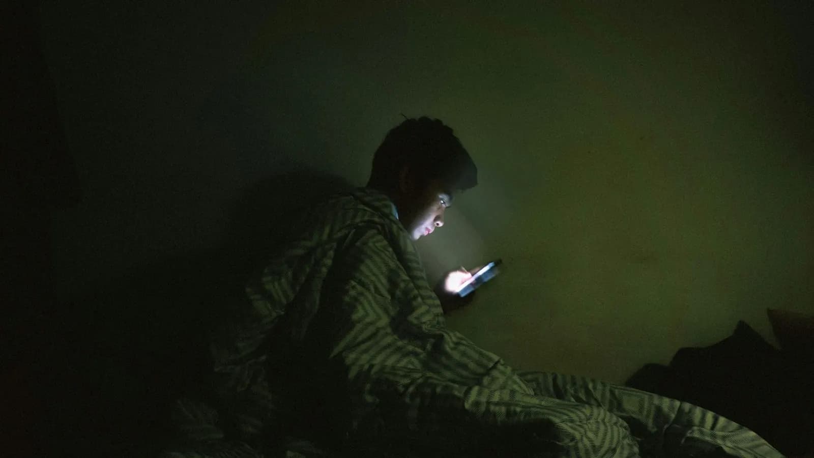 A young man in bed at night under a blanket, his face lit by his phone
