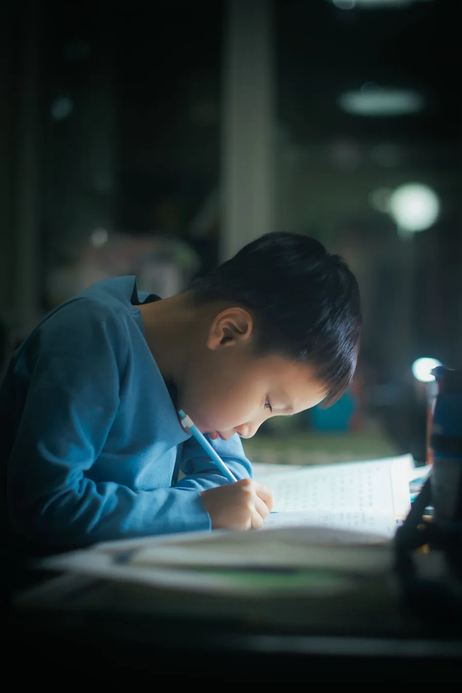 Desk lamp की रोशनी में अपनी copy में लिखता एक लड़का