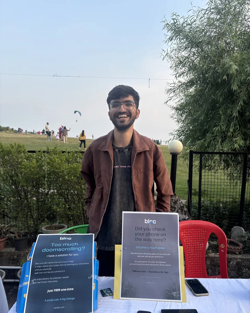 Smiling behind the bloc stall in Bir on a clear evening, bloc signs on the table and paragliders in the sky